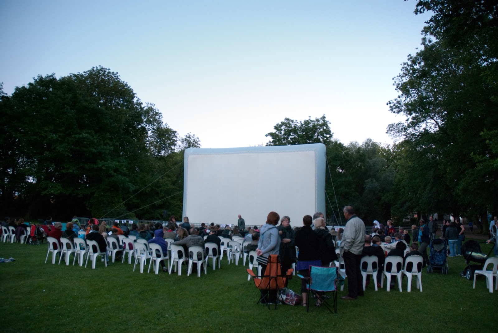Pantalla de cine hinchable gigante «Hallucine» instalada en un parque, con el público ya sentado al caer la tarde